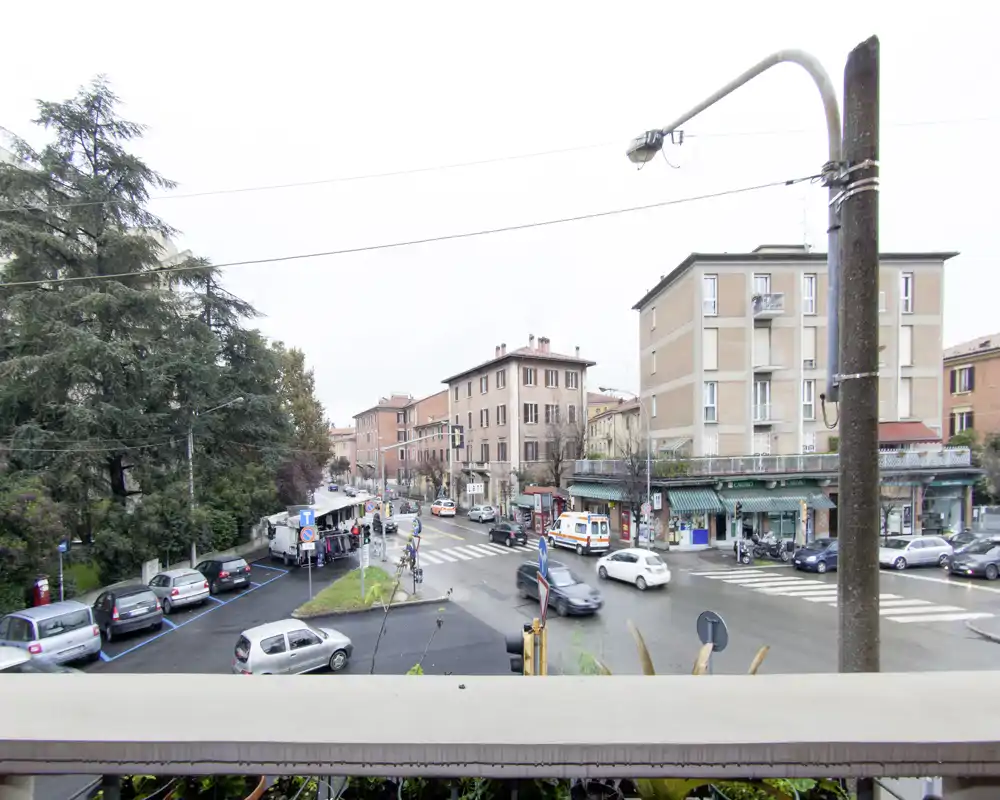 Veduta urbana da un balcone su un incrocio con auto parcheggiate, negozi e alberi; l'asfalto bagnato suggerisce pioggia recente.