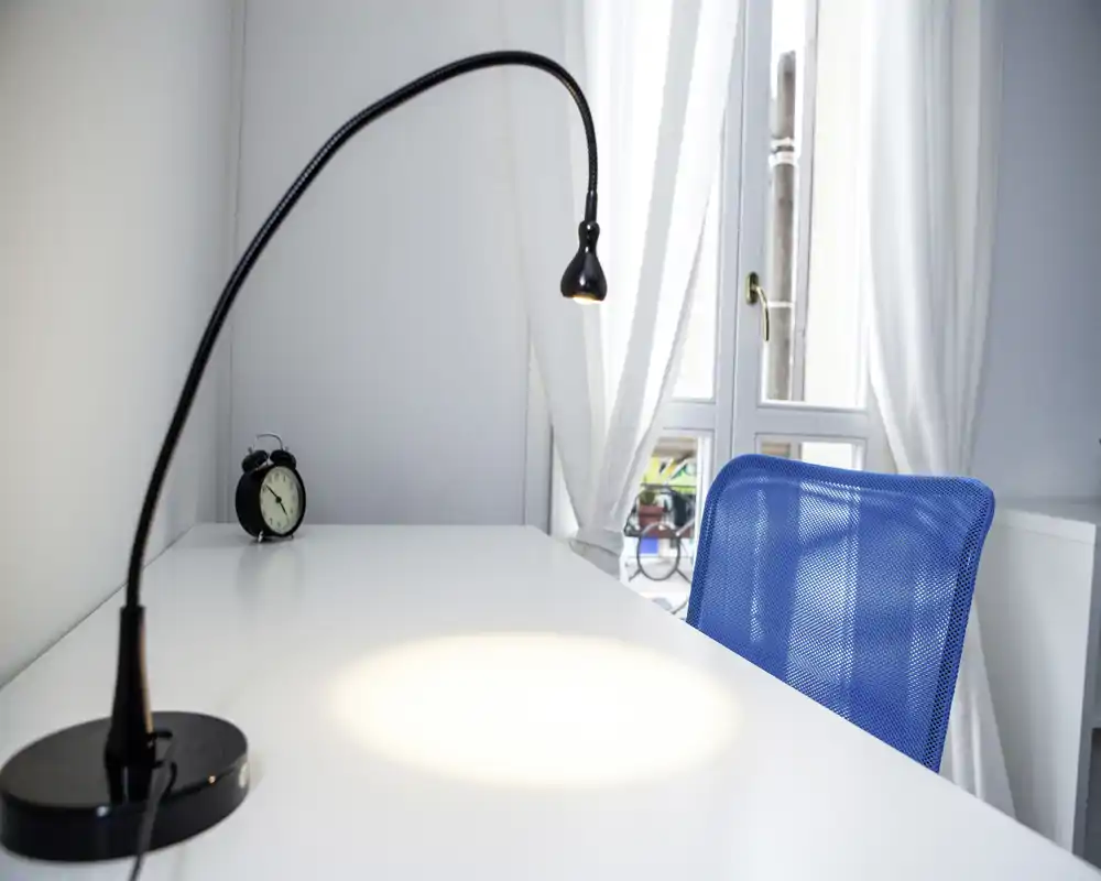 Angolo scrivania luminoso e minimale con lampada nera flessibile, sedia in rete blu e piano bianco accanto a una finestra con tende leggere. Perfetto per lavoro o lettura in un angolo compatto e arioso.