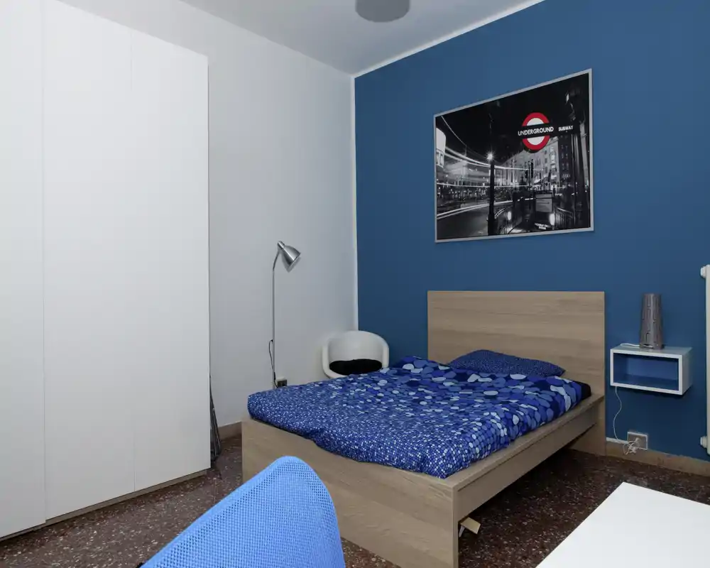 Camera da letto moderna e pulita con letto matrimoniale con biancheria blu, testiera in legno, armadio e arredi minimal; luminosa e ben curata, ideale per annunci.