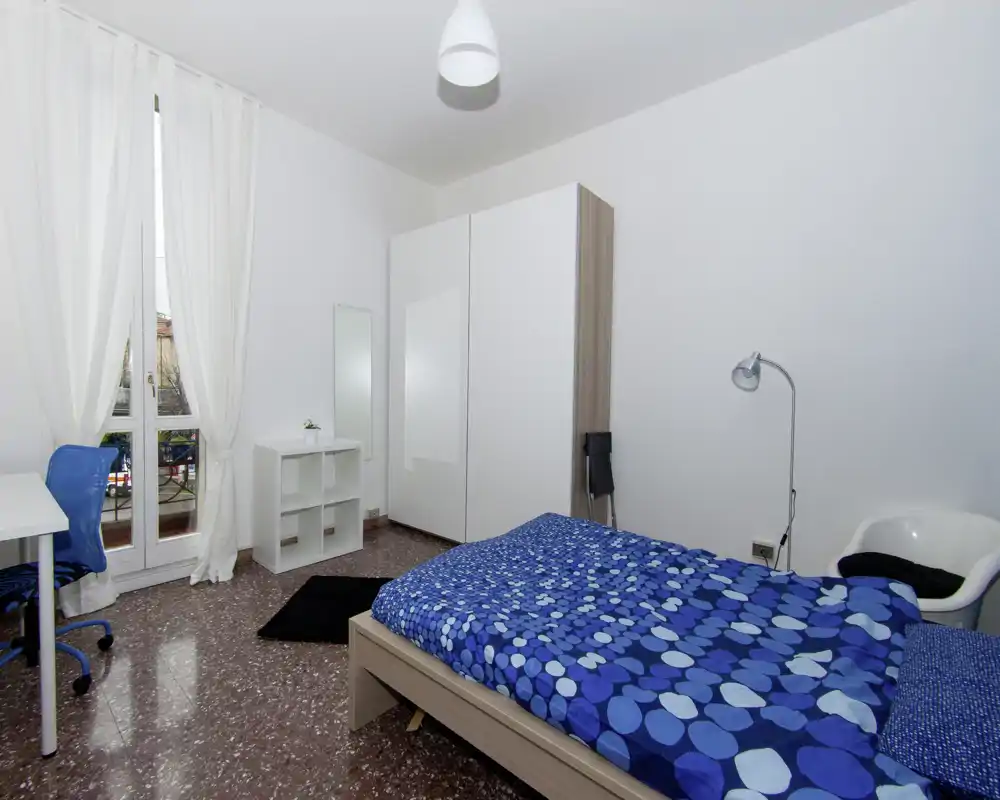 Camera da letto luminosa e ordinata con letto matrimoniale con copriletto blu a fantasia, armadio, scrivania e finestra con balcone che illumina la stanza.