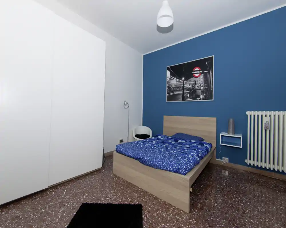 Camera da letto moderna e luminosa con letto matrimoniale in legno su parete accent color turchese. Armadio bianco capiente, lampada e arredi essenziali su pavimento in graniglia, accogliente e ben illuminata.