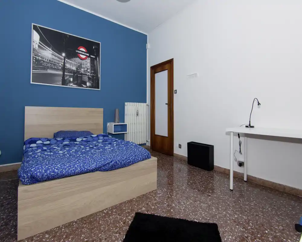Camera da letto luminosa e ordinata con struttura letto in legno, parete blu accent e scrivania semplice — ambiente confortevole e ben illuminato ideale per una persona.