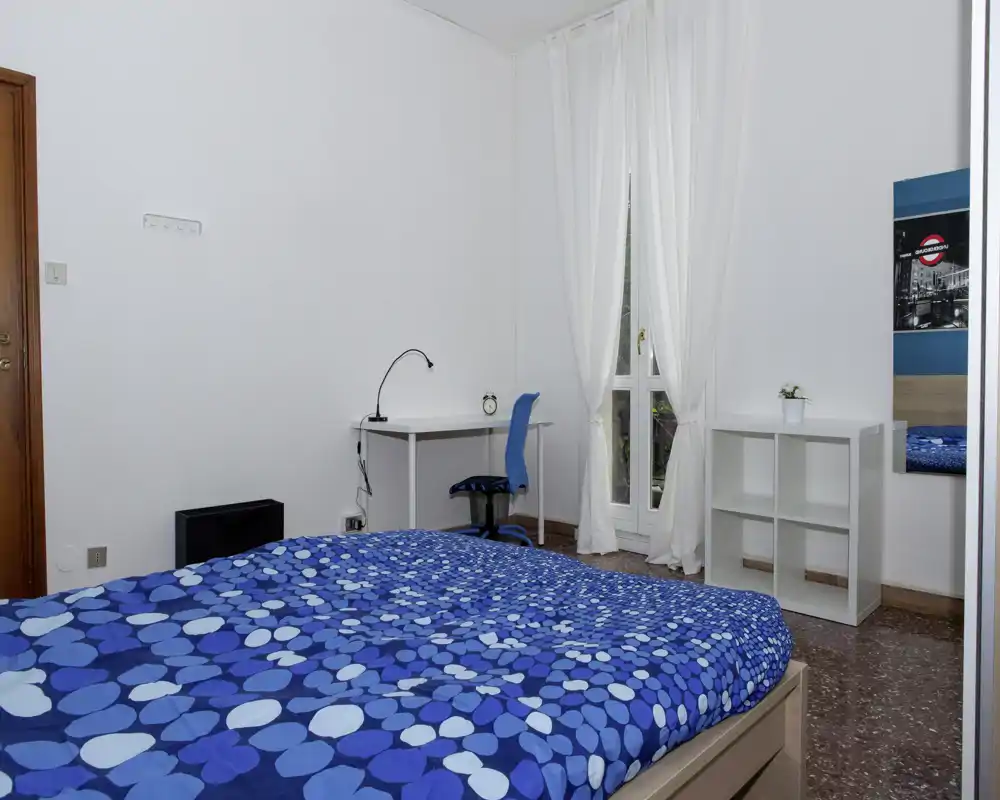 Camera da letto luminosa e ordinata con un ampio letto con copriletto blu a fantasia, una scrivania con sedia vicino a una finestra alta e scaffalatura semplice — ideale per una persona o una coppia.