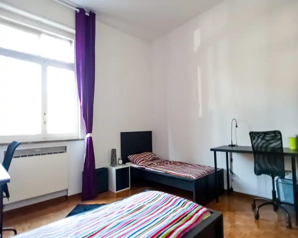 Bright, tidy bedroom with two single beds, striped bedding and a work desk by the wall. Large window provides abundant natural light and simple, modern furnishings create a comfortable stay.