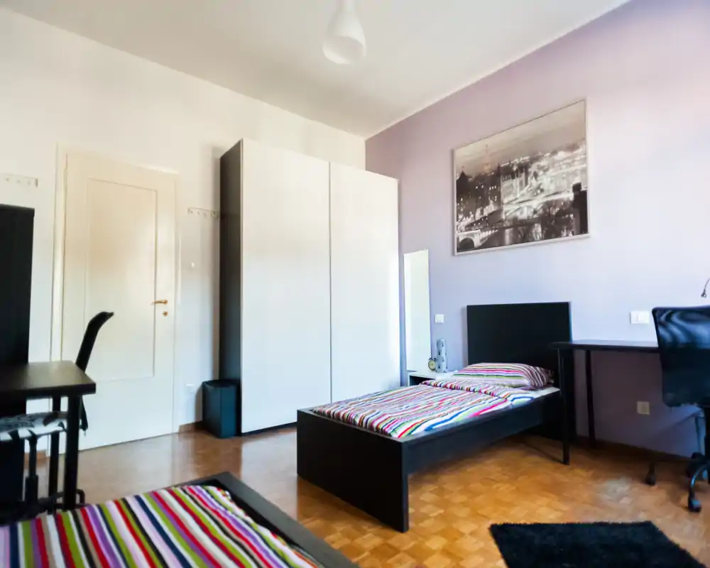 Bright, modern bedroom with a single bed, desk and wardrobe; colorful striped bedding and a framed city photo create a tidy, welcoming space ideal for a student or short-term stay.