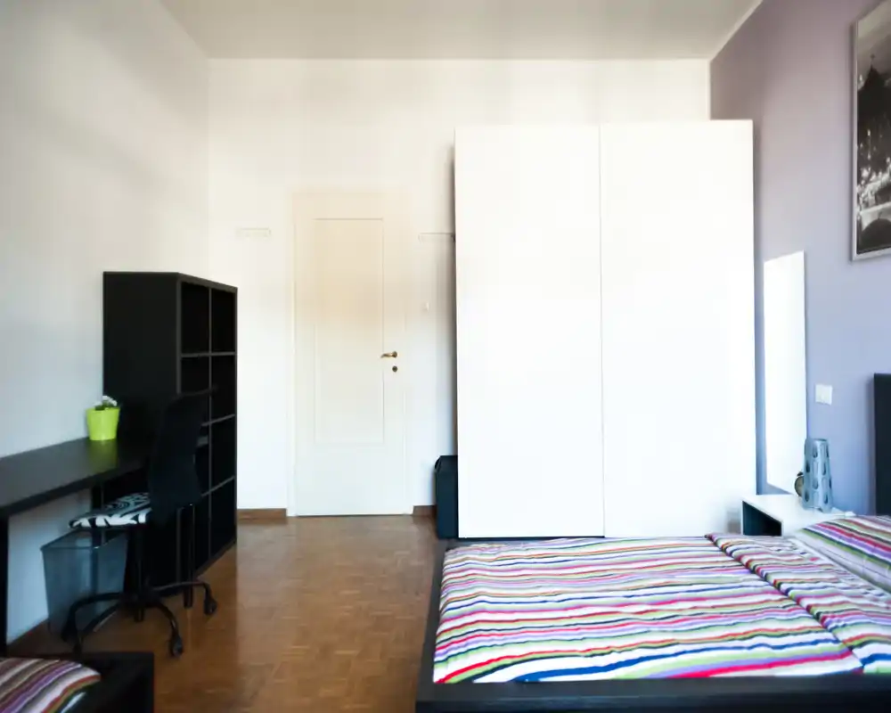 Bright, tidy bedroom with a double bed dressed in colorful striped bedding, a large white wardrobe and a desk area — ideal for a comfortable short or long stay.