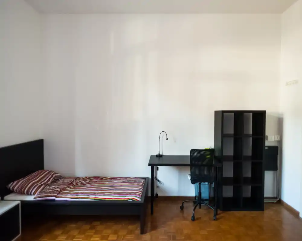Minimal, well-lit bedroom with a single bed, striped bedding, a desk and shelving unit—clean, functional layout ideal for a student or single occupant.