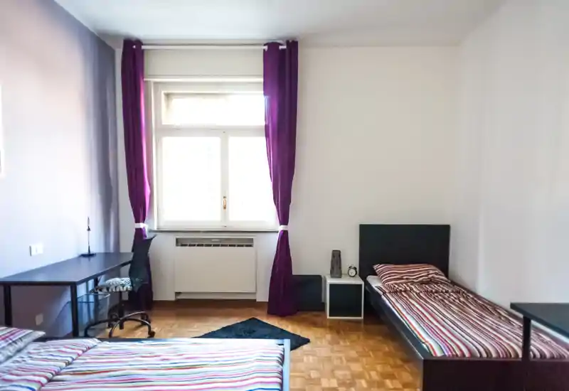 Bright, simple bedroom with two single beds, striped bedding and a desk by the window; purple curtains add a pop of color and natural light fills the room.