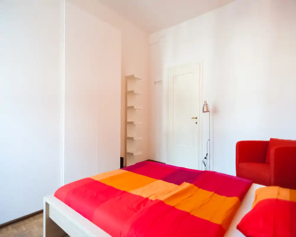 Bright, modern bedroom with a colorful double bed and a red armchair. Clean white walls and built-in wardrobe create a fresh, minimalist look.