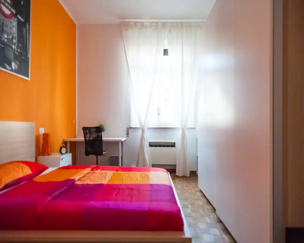 Bright, colorful bedroom with a double bed, bedside table and a small desk by the window. Natural light and warm orange accent wall create a welcoming feel ideal for rental listings.