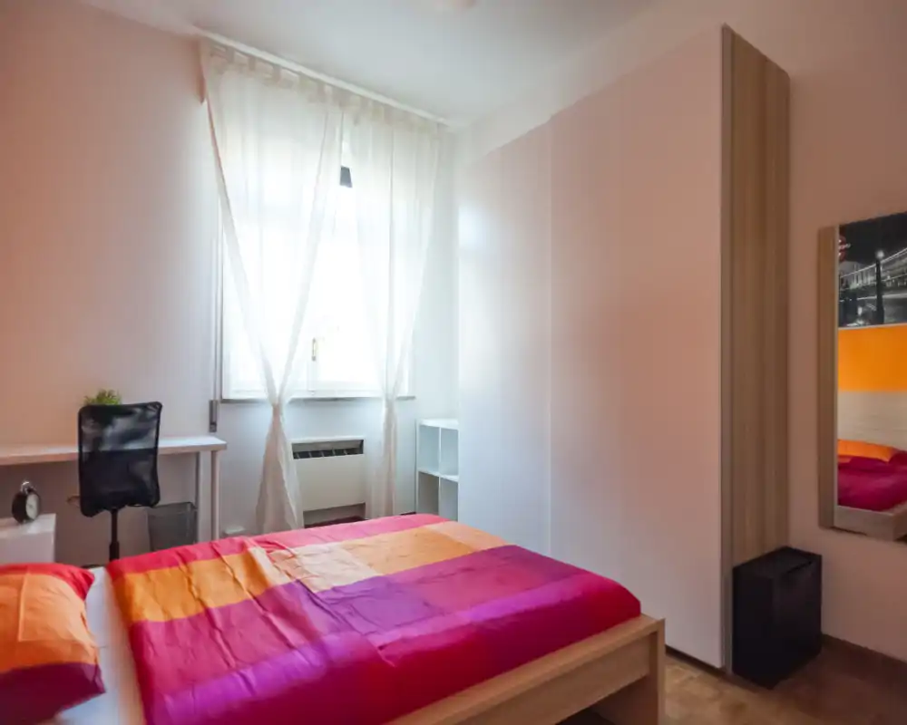 Bright, cozy bedroom with a colorful double bed, large wardrobe and natural light from a central window; a small desk and mirror complete the functional layout.