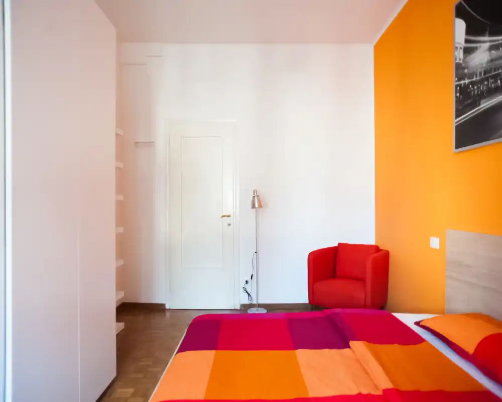 Bright, colorful bedroom with a large bed dressed in warm orange and red linens, a cozy red armchair and minimalist décor offering a cheerful, contemporary feel.