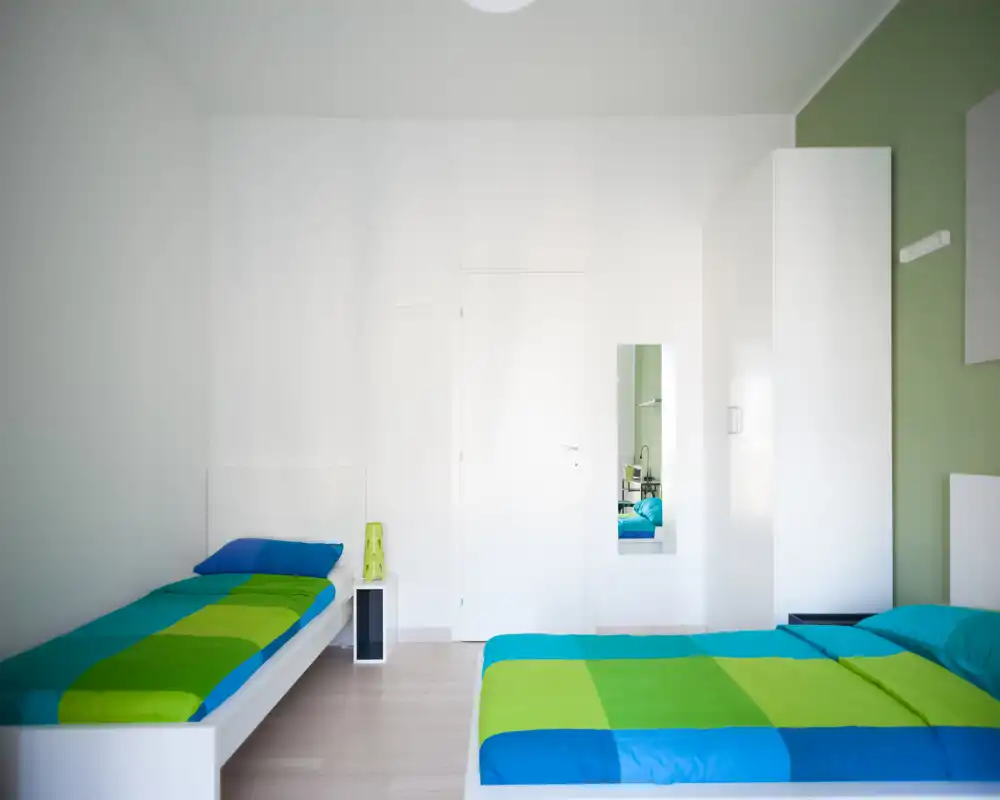 Bright modern bedroom with two single beds dressed in vivid blue and green bedding, a white wardrobe and minimal bedside furniture. Clean, airy space ideal for comfortable short-term stays.