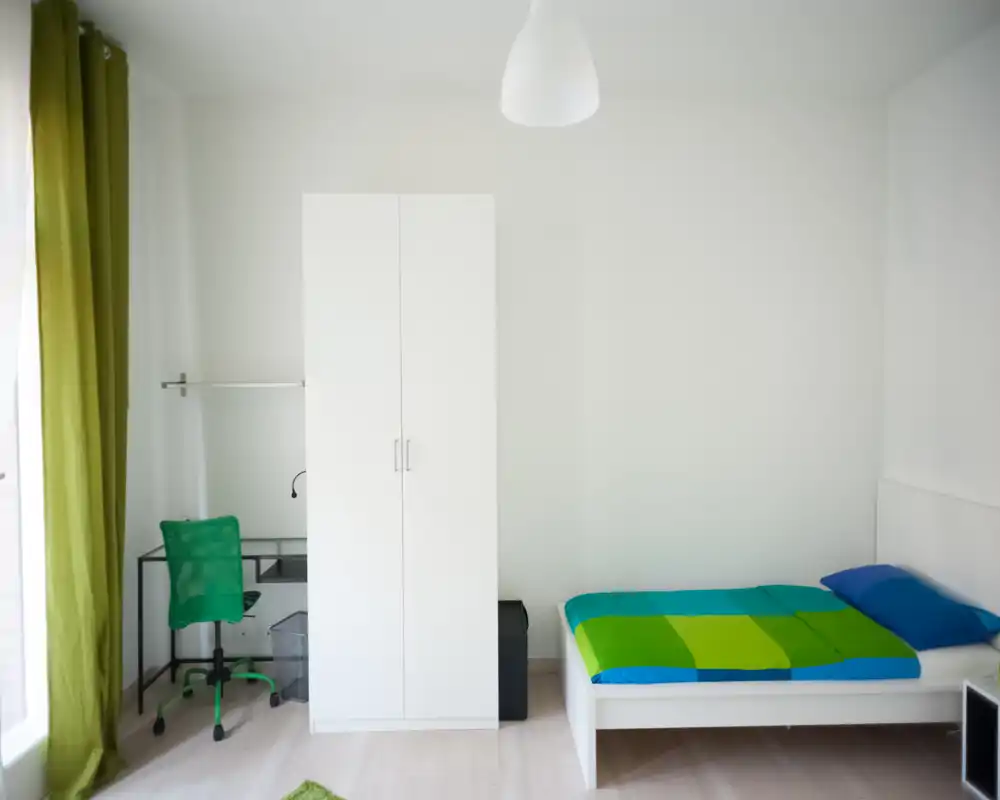 Bright, minimal bedroom with a single bed dressed in a vibrant green-and-blue duvet, a white wardrobe and a compact desk with a green chair beside a large window.