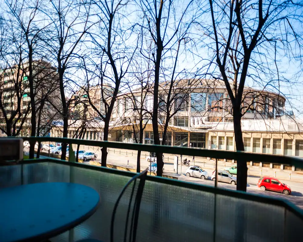 Sunny balcony view overlooking a tree-lined street and a distinctive circular building; small outdoor table and chairs in the foreground create a cozy spot to relax.
