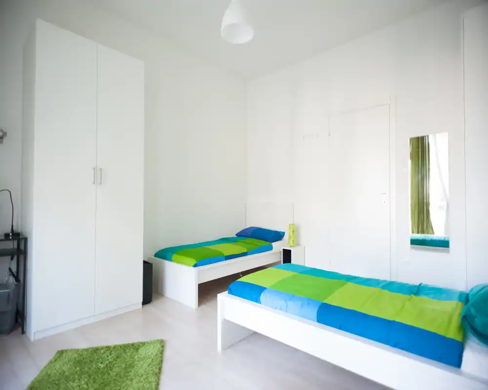 Bright, minimal twin bedroom with two single beds, colorful blue-green bedding and a simple wardrobe. Clean, airy composition ideal for short-term rentals or guest rooms.