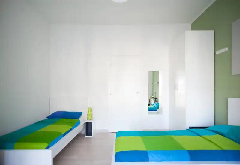Bright modern bedroom with two single beds dressed in vivid blue and green bedding, a white wardrobe and minimal bedside furniture. Clean, airy space ideal for comfortable short-term stays.