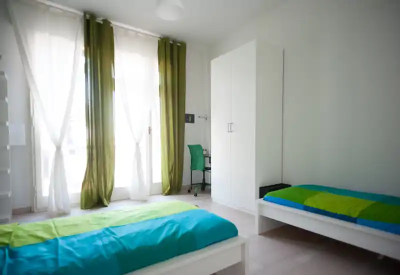 Bright twin-bedroom with two single beds, large windows with sheer and green curtains, a wardrobe and a small desk—fresh, airy and well-lit.