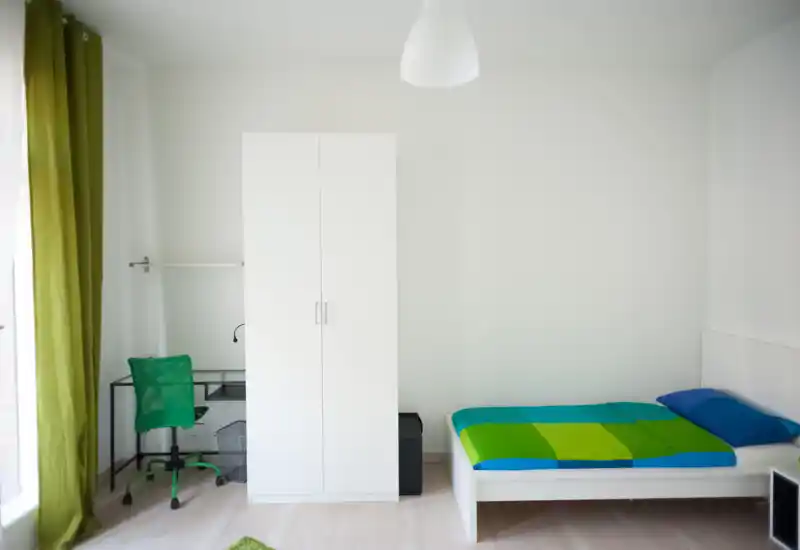 Bright, minimal bedroom with a single bed dressed in a vibrant green-and-blue duvet, a white wardrobe and a compact desk with a green chair beside a large window.