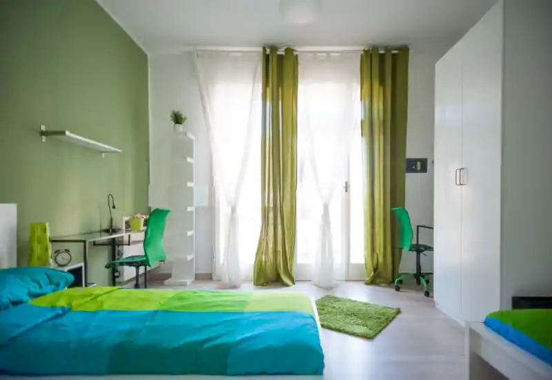 Bright, modern bedroom with a large bed dressed in turquoise and green linens, two study areas, and a sunlit balcony framed by sheer curtains and green drapes.