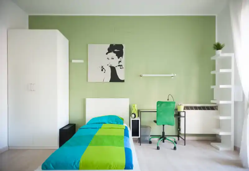 Bright, modern single bedroom with a colorful green-blue bed, a compact desk and shelving. The room shows clean lines, natural light from the window and tasteful wall art—ideal for a student or single occupant.