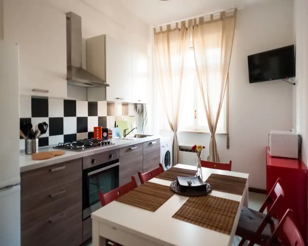 Bright modern kitchen with a gas stove, oven, ample counter space and a dining table for four. Light curtains and decorative placemats create a cozy, inviting atmosphere.