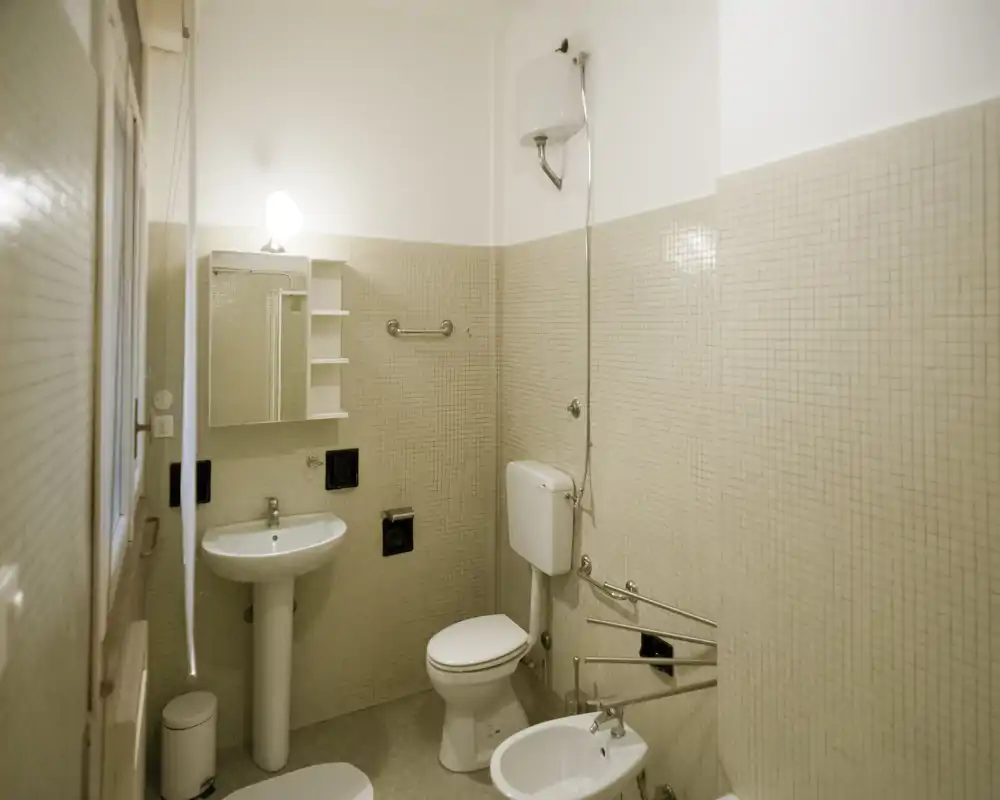 Compact, well-lit bathroom with sink, mirror cabinet, toilet and bidet; neutral mosaic tiles and simple fixtures suitable for rental listings.