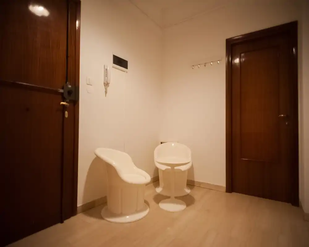 Small entrance hallway with two modern white chairs, wooden doors and a wall-mounted intercom; neutral lighting creates a warm, welcoming atmosphere.