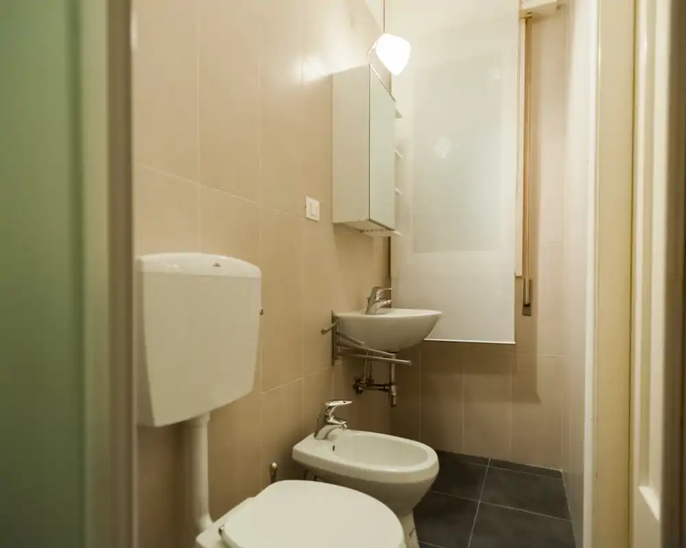 Compact, well-lit bathroom with toilet, bidet and wall-mounted sink. Neutral tiles and a frosted window blind create a clean, functional space.