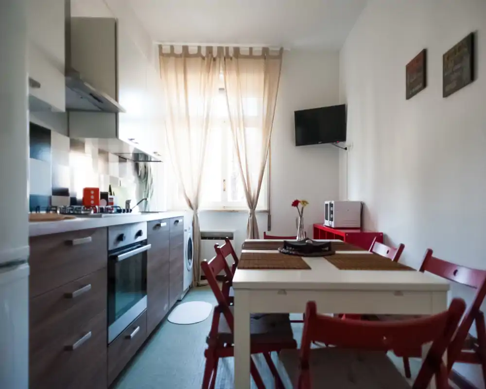 Bright, well-equipped kitchen with modern cabinets, gas hob and an inviting dining table with red chairs—ideal for shared meals. Natural light filters through soft curtains creating a warm, homely atmosphere.