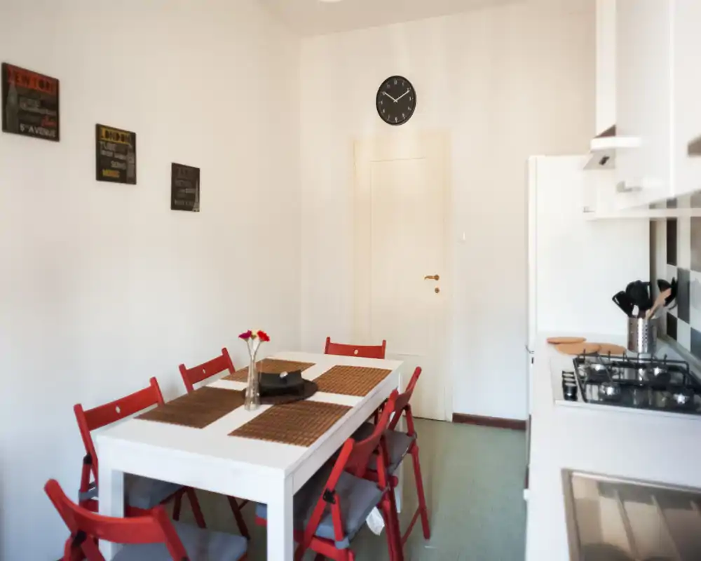 Bright, compact kitchen-dining area with a white table, red folding chairs and a gas hob along the right wall; simple décor with placemats and a small vase of flowers.