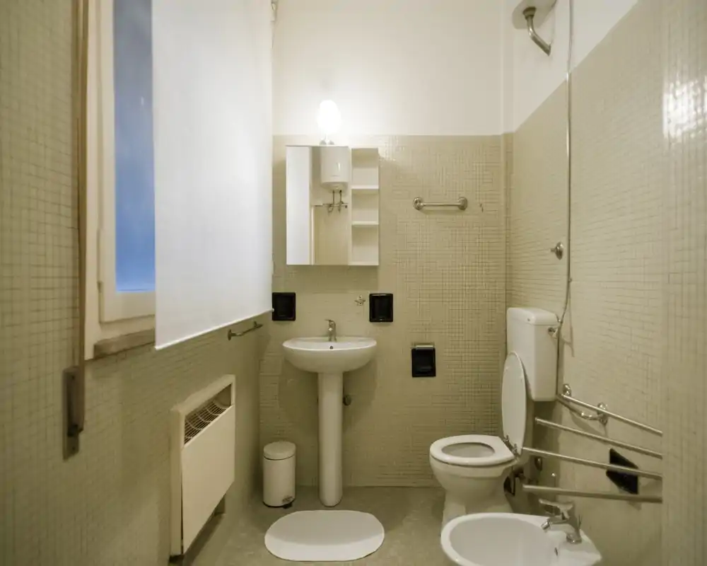 Clean, compact bathroom with a pedestal sink, toilet, bidet and wall-mounted cabinet; soft neutral tiles and a window provide natural light.