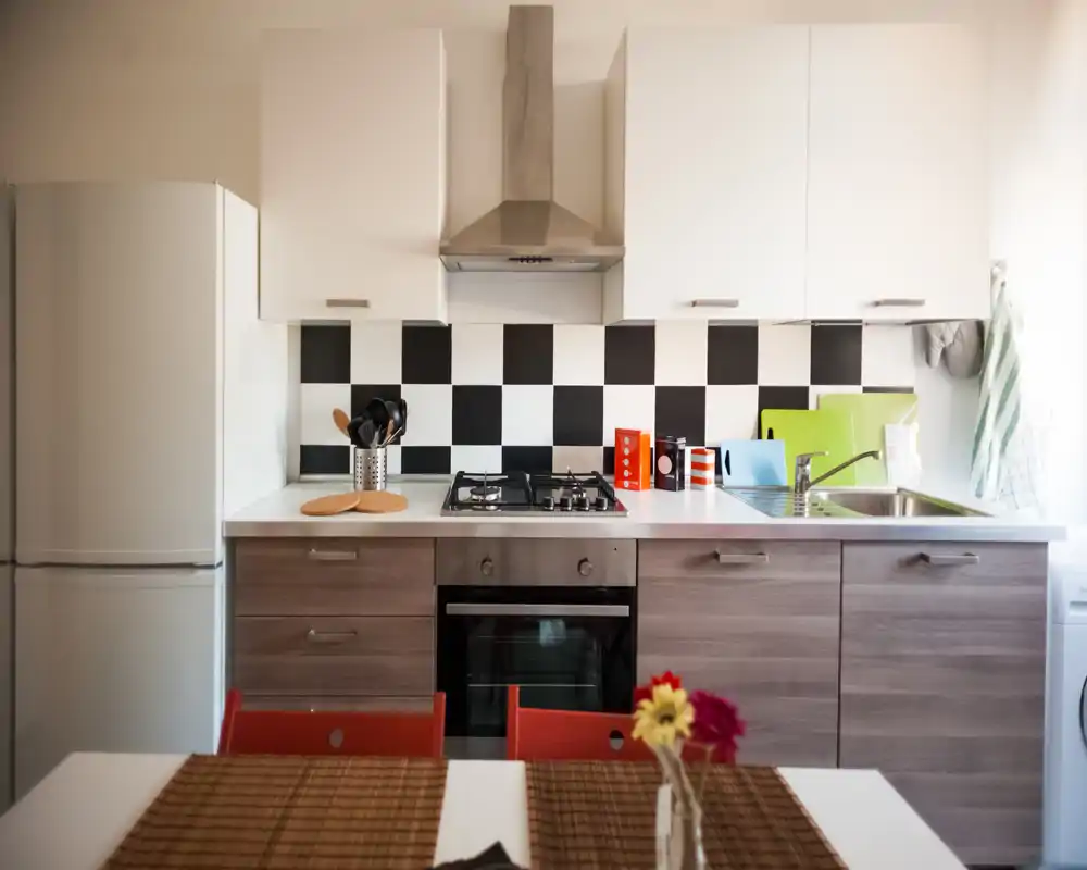 Bright, tidy modern kitchen with a checkerboard backsplash, gas hob, built-in oven and fridge. A small dining surface with placemats and fresh flowers adds a welcoming touch.