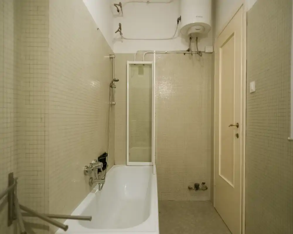 Compact bathroom with a bathtub and wall-mounted showerhead, tiled walls and a water heater mounted high on the wall. Neutral tones and simple fixtures.