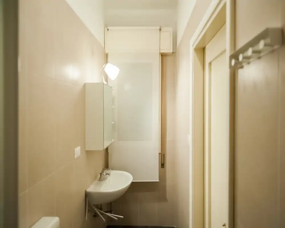 Clean, compact bathroom with a wall-mounted sink, mirrored cabinet and soft warm lighting. Neutral tiles and a roller blind create a minimalist, functional look ideal for a rental listing.