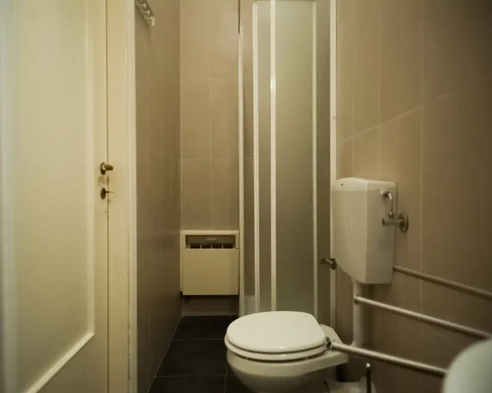 Compact tiled bathroom with a wall-mounted toilet and a narrow shower cubicle; neutral beige tiles and modest lighting create a clean, functional space.