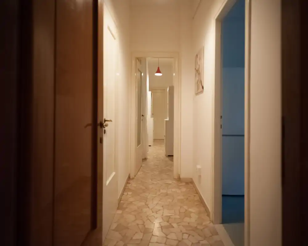Narrow indoor hallway with tiled floor, white walls and several doorways leading to other rooms; softly lit with a pendant light visible in the distance.