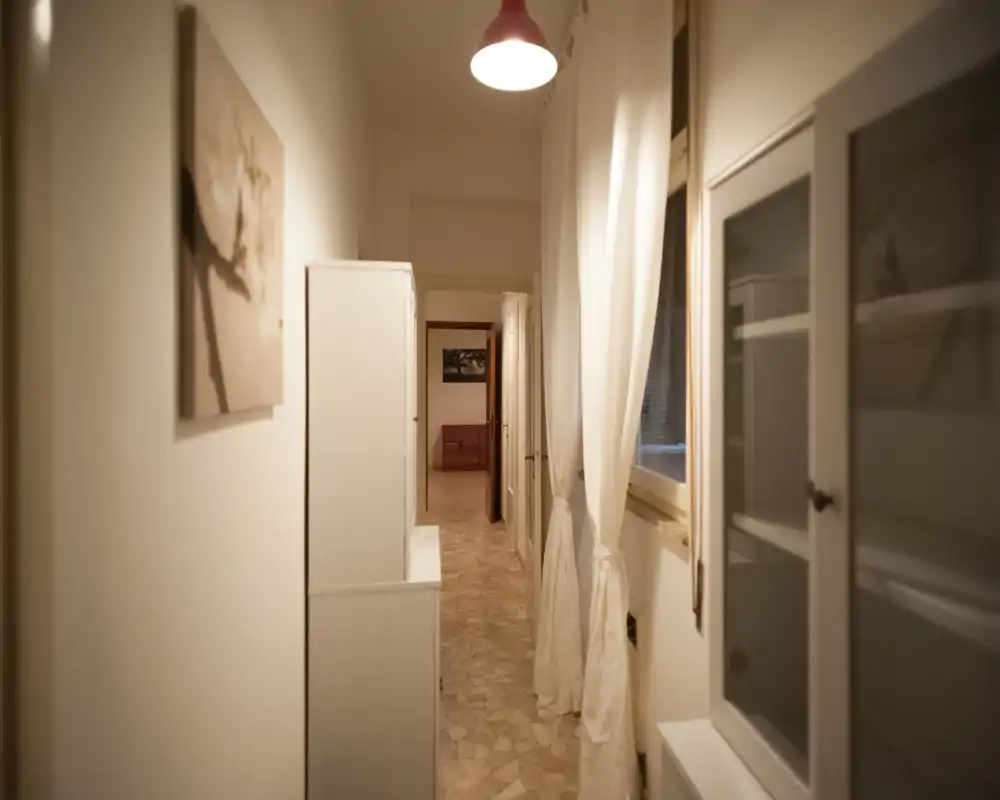 Narrow, well-kept hallway with tiled floor, soft warm lighting and simple white furnishings — ideal as an entrance corridor connecting rooms.