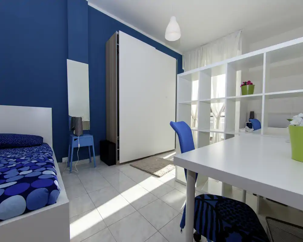 Camera da letto moderna e luminosa con parete blu, letto singolo con copriletto a fantasia blu, scrivania e scaffalatura che creano un'area studio funzionale e ben illuminata.