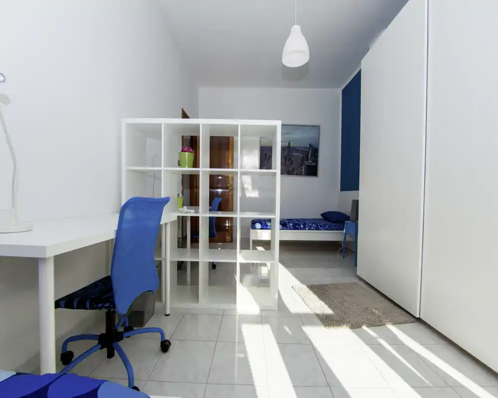 Camera da letto luminosa e ordinata con un letto singolo, armadio a muro e una scrivania separata da scaffalatura aperta; pareti bianche e dettagli blu per un aspetto moderno e arioso.