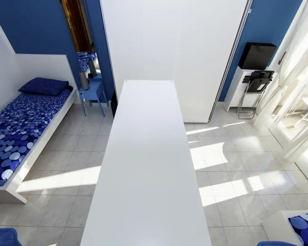 Camera singola luminosa e moderna con biancheria blu, pavimento in piastrelle bianche e molta luce naturale dalla porta del balcone. Armadio e arredo semplice completano lo spazio, perfetto per un affitto compatto e di stile.