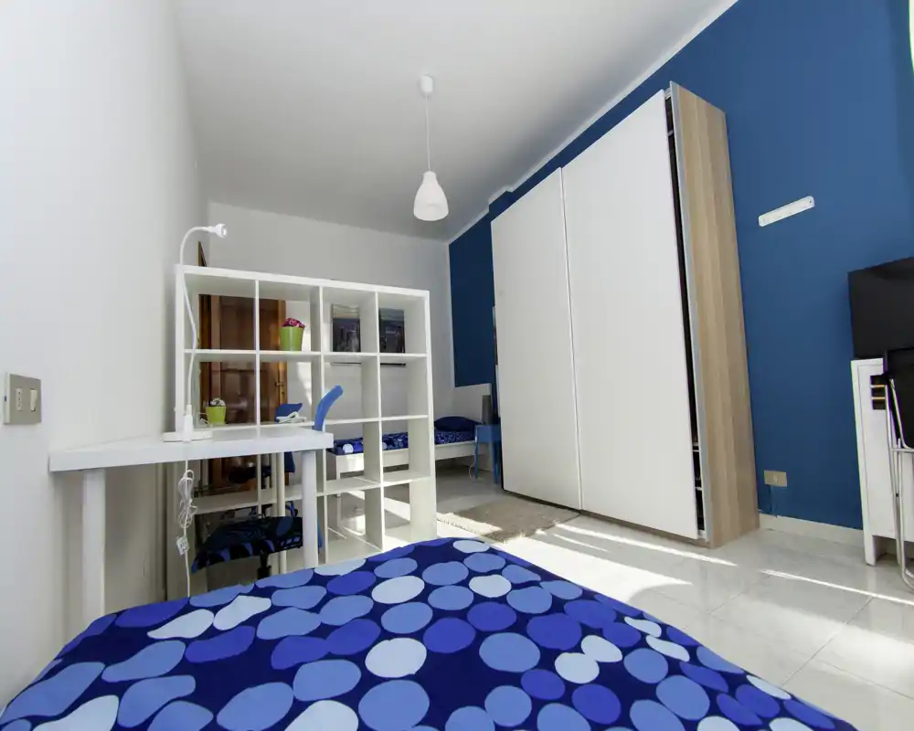 Camera da letto moderna e luminosa con copriletto blu a pois, un divisorio a scaffali bianco e un ampio armadio su una parete blu intensa. La stanza è ben illuminata dalla luce naturale e arredata in modo ordinato.
