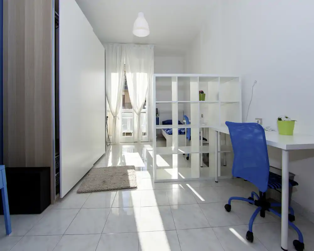 Camera da letto luminosa e moderna con il letto visibile dietro una libreria bianca a giorno, una scrivania con sedia blu e molta luce naturale da una grande finestra.