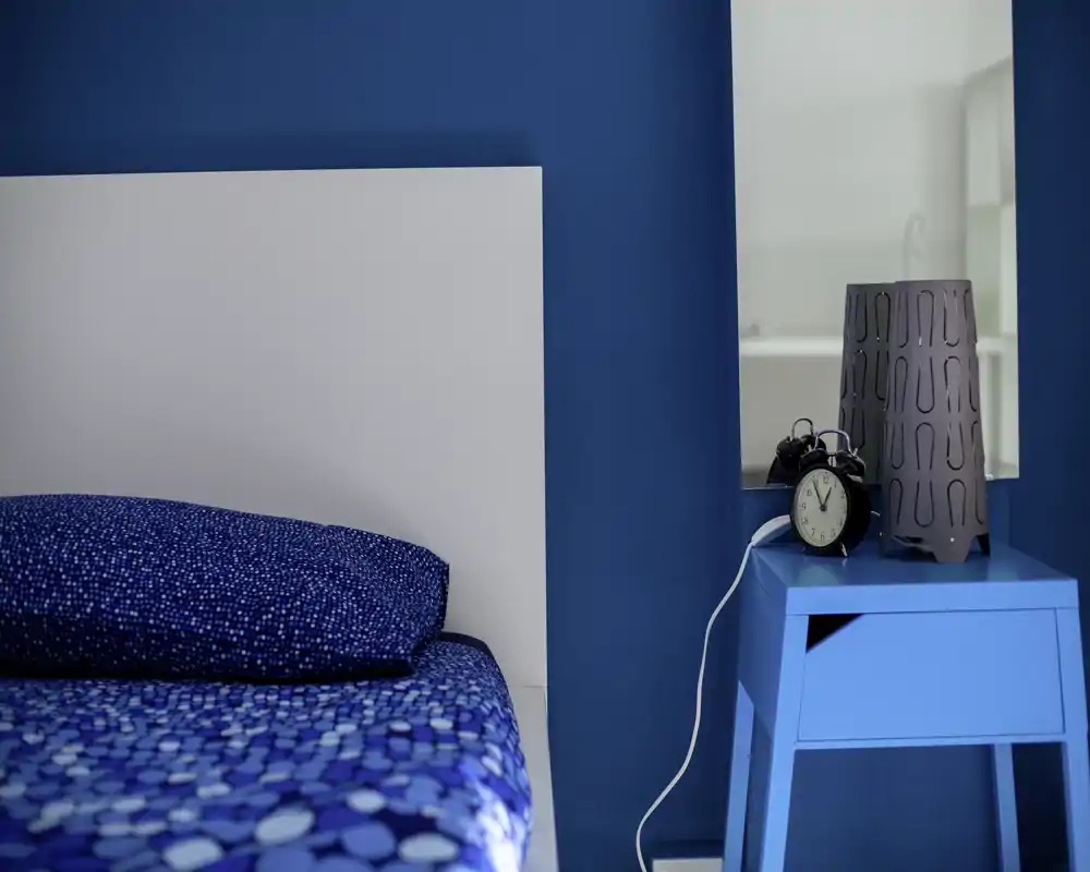 Angolo di camera luminoso e moderno con copriletto blu a motivi e una testiera bianca; un piccolo comodino blu ospita una sveglia vintage e una lampada decorativa.