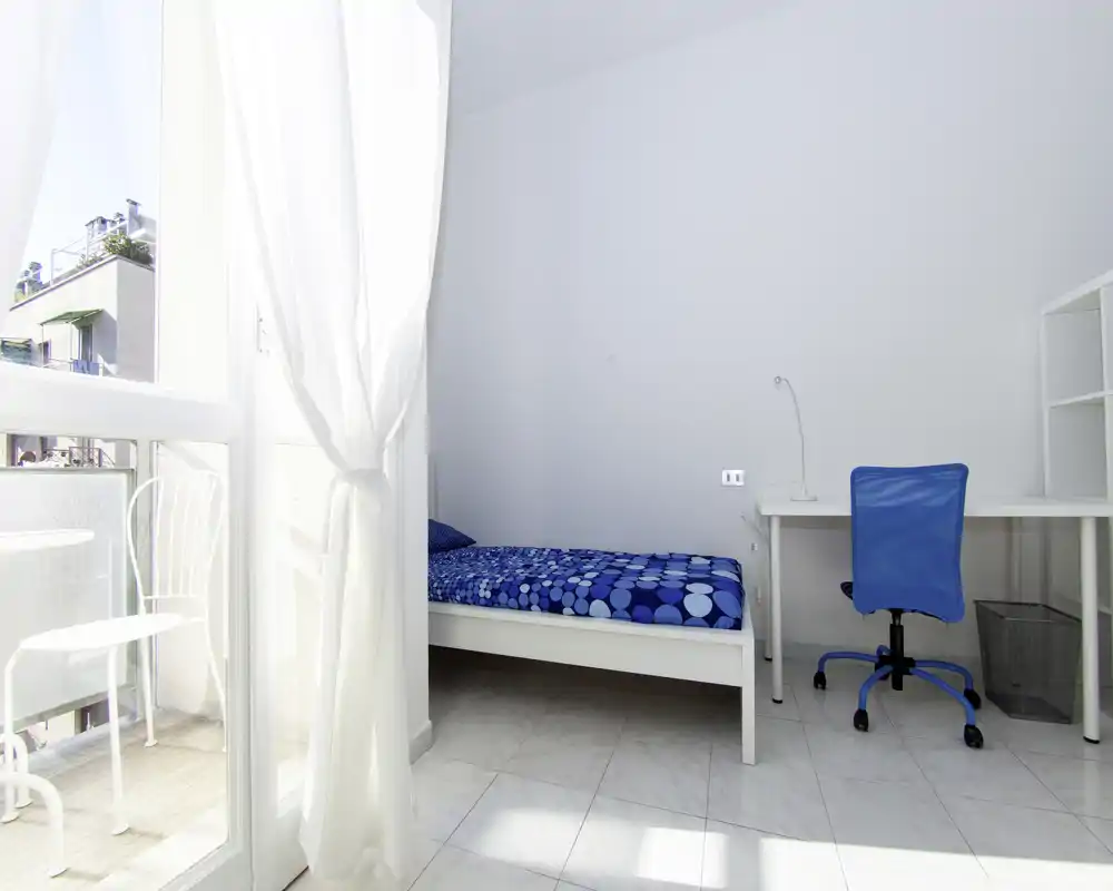 Camera singola luminosa e minimale con piumone blu a motivi, scrivania e accesso al balcone. Pareti bianche e luce naturale abbondante la rendono ideale per studenti o affitti brevi.