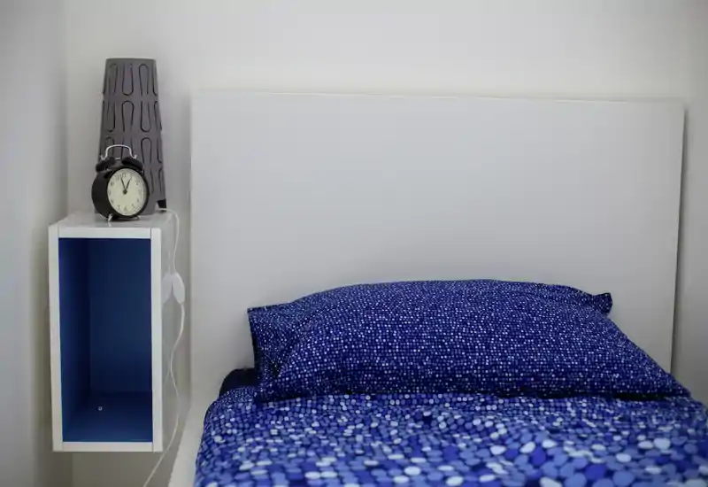 Bright, minimal bedroom close-up showing a neatly made bed with blue patterned bedding and a simple white headboard; a bedside shelf with a lamp and alarm clock is visible.