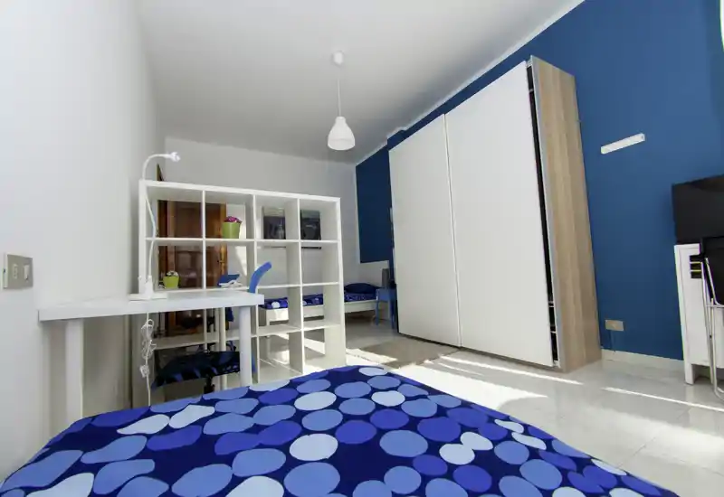 Bright, modern bedroom with patterned blue bedding, a white shelving divider and a large wardrobe against a deep blue accent wall. The room is well lit with natural light and neatly staged for a comfortable stay.
