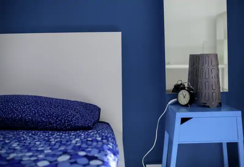 Bright, modern bedroom corner with a patterned blue duvet and a white headboard; a small blue bedside table holds a vintage alarm clock and a decorative lamp.