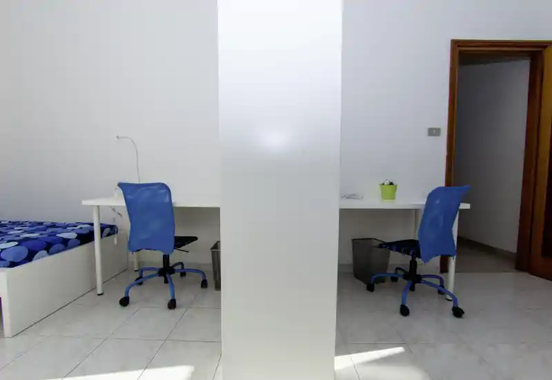 Bright, minimal bedroom featuring a single bed with blue patterned bedding and two work desks with blue chairs separated by a central partition. Clean, modern styling and abundant natural light make it suitable for a student or shared flat.