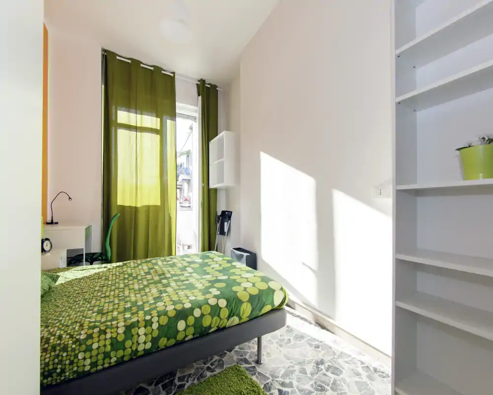 Chambre lumineuse et soignée avec une couette verte à motifs, lumière naturelle venant d'une fenêtre avec rideaux verts et étagères minimalistes. L'espace paraît frais et accueillant, parfait pour une annonce locative.